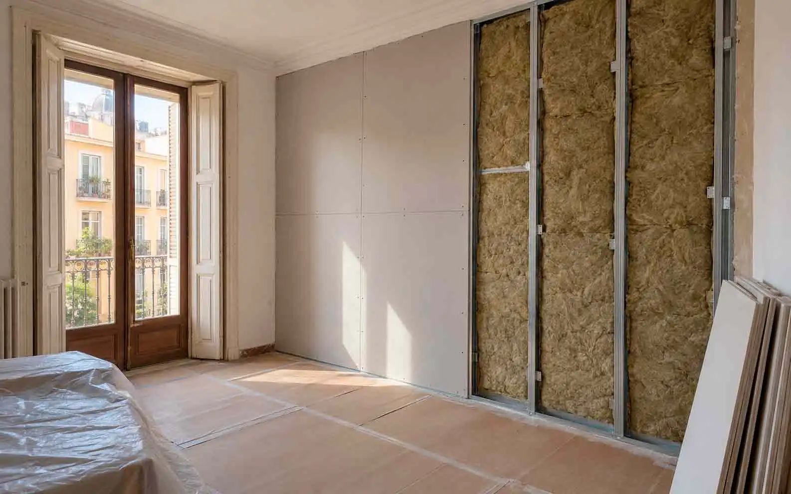 Dormitorio con balcón en plena obra: parte de la pared ya cerrada con placas de yeso y el resto con lana mineral entre perfiles metálicos, con el suelo y la cama protegidos