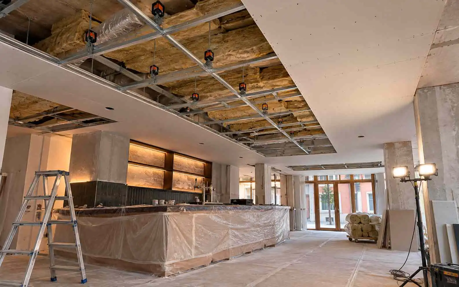 Bar en obra con el techo abierto: estructura de perfiles colgada con amortiguadores y lana mineral, la barra tapada con plástico, una escalera y focos de trabajo
